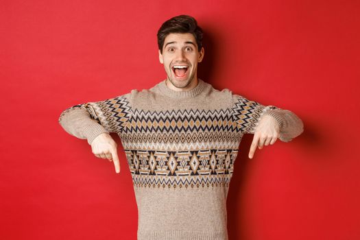 Concept of christmas celebration, winter holidays and lifestyle. Excited caucasian male model in xmas sweater, pointing fingers down and making announcement, showing advertisement, red background.