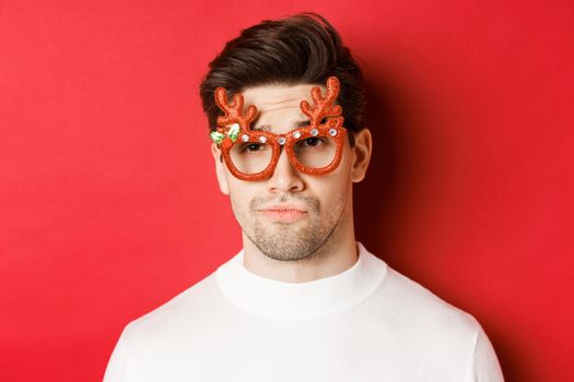 Concept of winter holidays, christmas and celebration. Close-up of skeptical handsome guy, wearing party glasses, smirk and looking unamused, standing over red background.