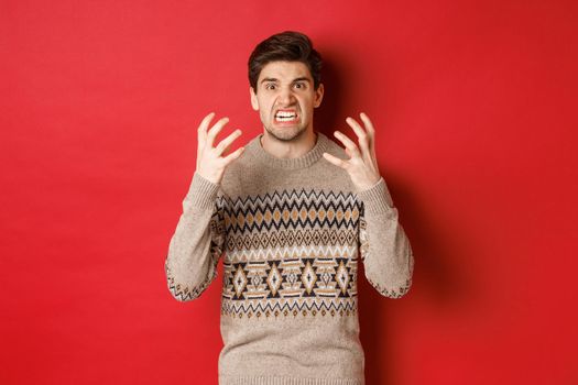 Image of angry and frustrated man in winter sweater, shouting and looking with hatred, standing upset over red background.