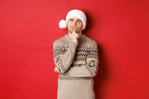 Image of attractive man in christmas sweater and santa hat, thinking about new year gifts, looking at upper left corner thoughtful, standing over red background.