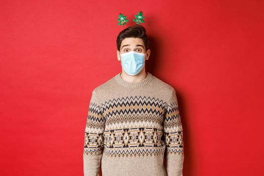 Concept of christmas, covid-19 and social distancing. Cheerful young man in medical mask and new year clothing, celebrating holidays during pandemic, standing over red background.