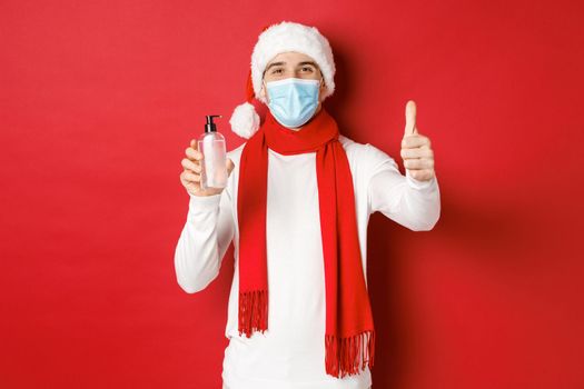 Concept of covid-19, christmas and holidays during pandemic. Happy and satisfied man in santa hat and medical mask, showing thumb-up in approval and recommending hand sanitizer.