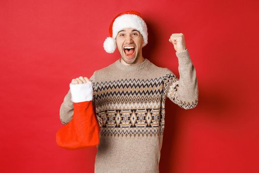 Concept of winter holidays, new year and celebration. Amazed and happy man shouting for joy, found gift inside christmas stocking and cheering, raising hand up and smiling.