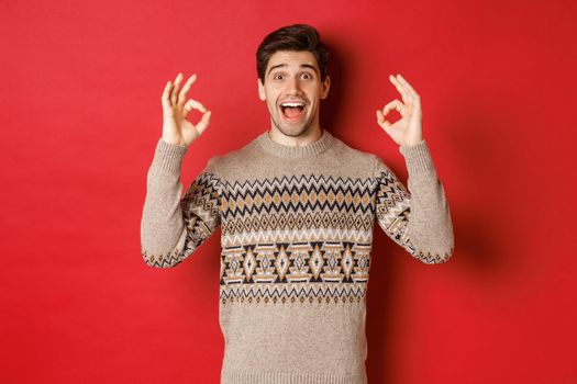 Concept of christmas celebration, winter holidays and lifestyle. Portrait of handsome man in sweater, looking amazed and showing okay signs, guarantee or recommend something good, red background.