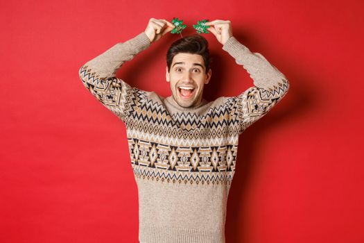 Concept of winter holidays, christmas and celebration. Image of funny and cute adult man enjoying new year party, smiling and rejoicing, standing over red background.