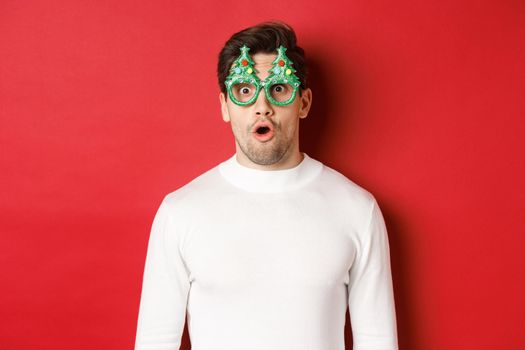 Concept of christmas, winter holidays and celebration. Close-up of surprised handsome guy in party glasses, saying wow and looking at amazing new year promo offer, red background.