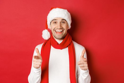 Concept of christmas, winter holidays and celebration. Cheeky man in santa hat and scarf, smiling and pointing fingers at camera, wishing happy new year, standing over red background.
