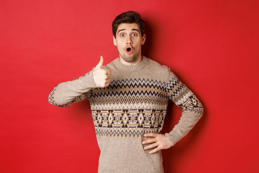 Image of handsome caucasian man in christmas sweater looking amazed, showing thumb-up in approval, like something, standing over red background.