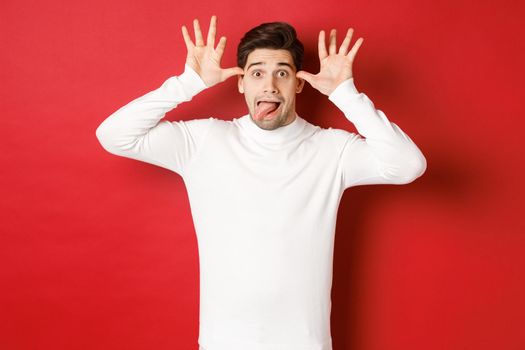 Portrait of funny caucasian guy, showing tongue and making faces, wearing white sweater, standing against red background.