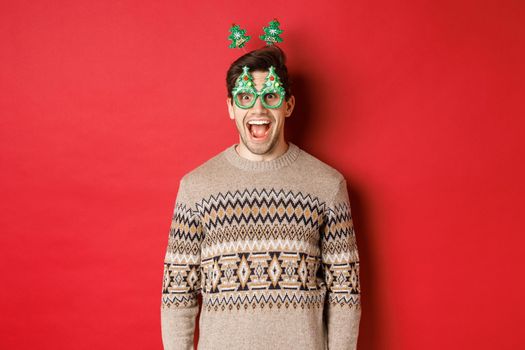 Portrait of surprised and excited handsome guy in party glasses and winter sweater, celebrating christmas and having fun, standing against red background.