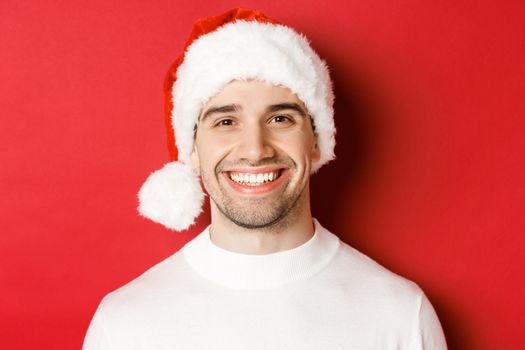 Close-up of attractive smiling man in white sweater and santa hat, looking happy, enjoying winter holidays, standing against red background.