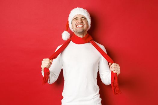 Image of distressed and frustrated man in santa hat, chocking himself with scarf from sadness, standing against red background.