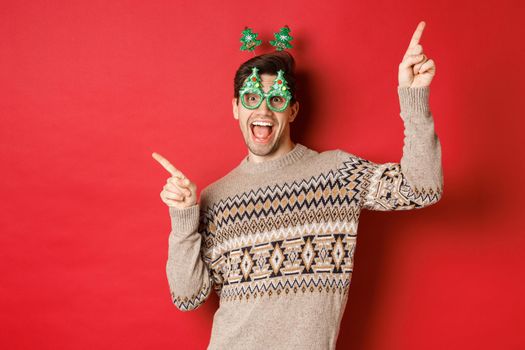 Portrait of joyful handsome guy in party glasses and christmas sweater, dancing and pointing fingers sideways, enjoying new year celebration, standing over red background.