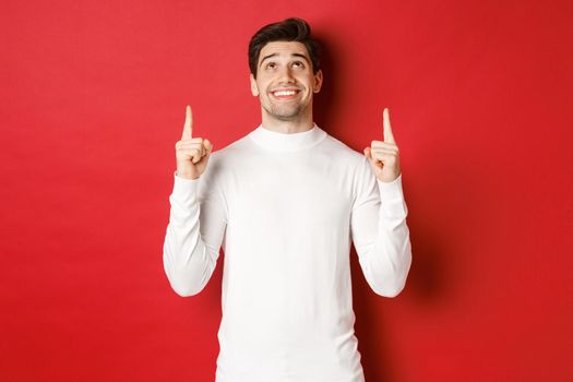 Concept of winter holidays. Image of happy attractive man in white sweater, smiling while pointing and looking up at copy space on red background.