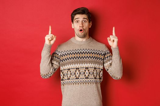 Portrait of surprised and amazed handsome guy showing christmas promo offer, wearing xmas sweater and standing over red background.