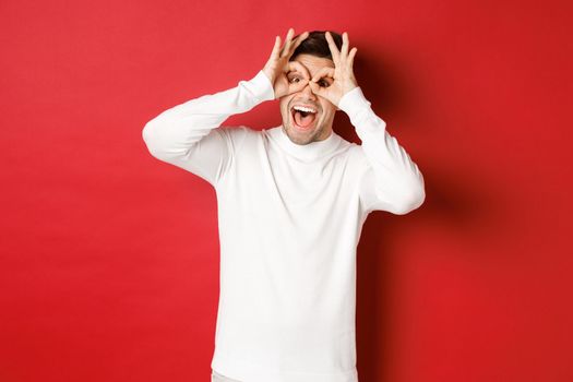 Portrait of handsome guy in white sweater, making funny mask with fingers, looking happy and smiling, standing over red background.