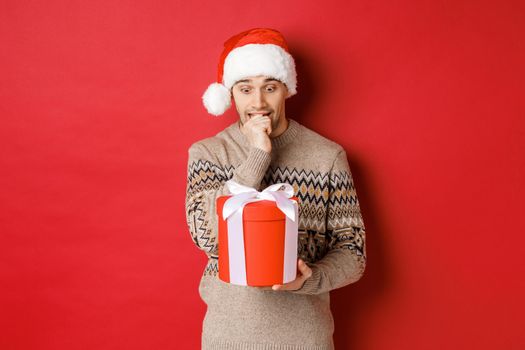 Image of worried guy in santa hat and sweater, looking indecisive at christmas gift, standing against red background.