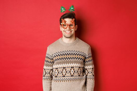 Portrait of handsome bearded guy in party glasses and winter sweater, smiling pleased, celebrating new year, wishing merry christmas, standing over red background.