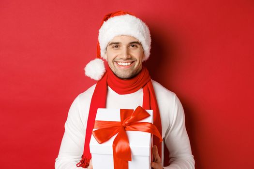 Concept of winter holidays, christmas and lifestyle. Close-up of attractive bearded guy in santa hat and scarf, holding a present, giving new year gift and smiling, red background.