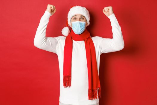 Concept of covid-19, christmas and holidays during pandemic. Portrait of happy man in santa hat and medical mask, rejoicing and celebrating new year, standing over red background.