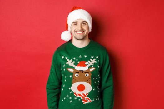 Concept of winter holidays, christmas and lifestyle. Handsome guy with bristle, wearing santa hat and green sweater, smiling joyful, celebrating new year, standing over red background.