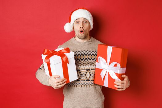 Concept of winter holidays, new year and celebration. Image of surprised attractive guy in santa hat and christmas sweater, receiving gifts, holding presents and looking amazed.