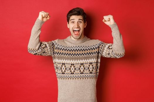 Image of happy handsome guy rejoicing on christmas eve, raising hands up and shouting for joy yes, achieve new year goal, standing over red background in sweater.