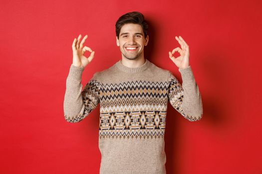 Concept of christmas celebration, winter holidays and lifestyle. Satisfied handsome man in sweater, showing okay signs and smiling pleased, recommending something, red background.