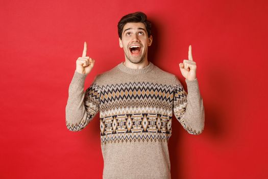 Image of excited handsome guy who likes winter holidays showing advertisement, looking and pointing fingers up with amazed face, standing in christmas sweater over red background.