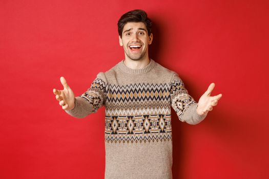 Image of happy and flattered handsome guy in christmas sweater, reaching hands to welcome guests on new year party, inviting to celebration, standing over red background.