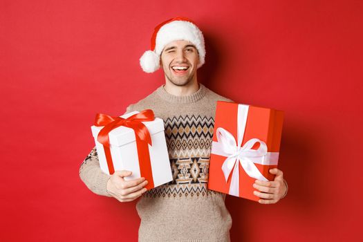 Concept of winter holidays, new year and celebration. Portrait of confident and cheeky young man prepared gifts for christmas, winking and holding presents, standing over red background.