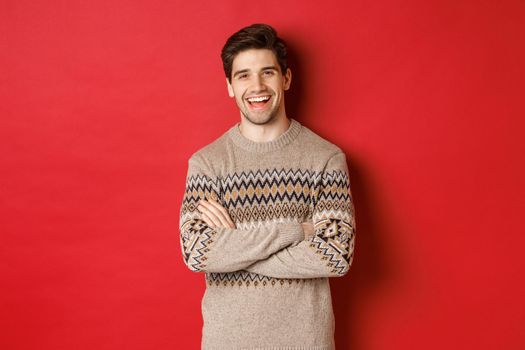 Portrait of cheerful, attractive man in christmas sweater, laughing and smiling, celebrating new year and winter holidays, standing over red background.