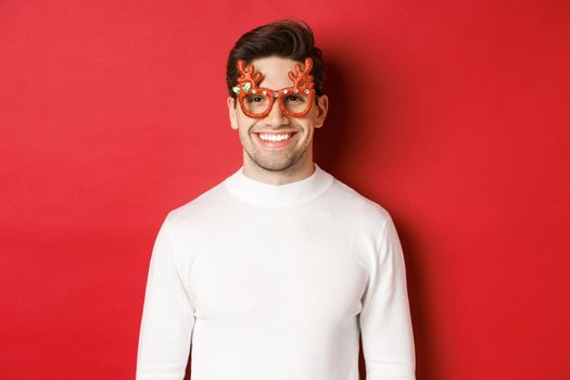 Close-up of attractive man in white sweater and party glasses, celebrating christmas, smiling amused, standing over red background.