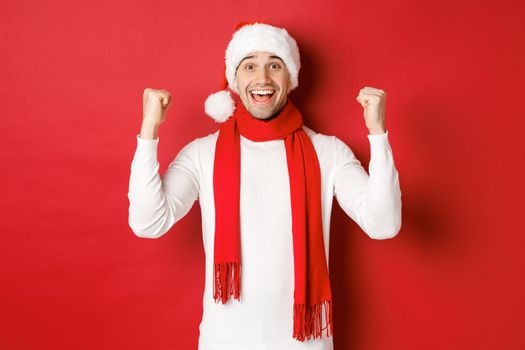 Portrait of happy and excited man in santa hat and scarf, rejoicing and winning something, celebrating new year, standing over red background.