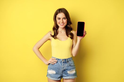 Stylish smiling woman showing mobile phone screen, app interface on smartphone, standing in tank top and shorts against yellow background.