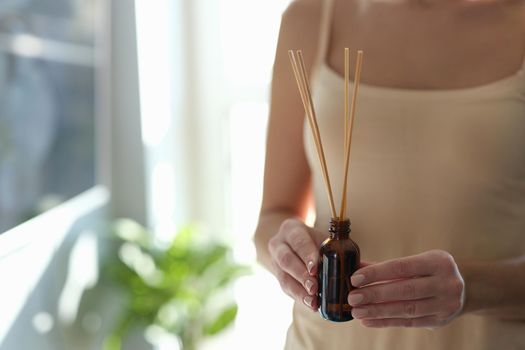Woman holding diffuser with bamboo sticks, sunny, close-up. Aromatherapy at home, perfume, relaxation spa