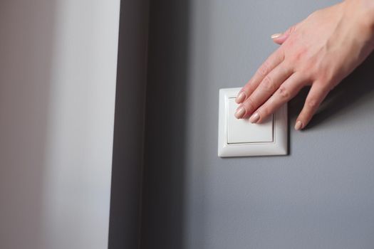 A woman's hand presses the switch on the wall, close-up. White light switch on gray wall background, checking electricity