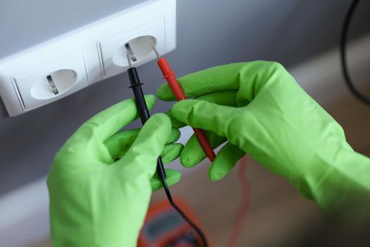 Hands in green gloves plug the voltmeter into an outlet, close-up. Home electrical wiring repair, electrician equipment