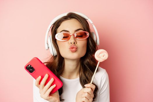 Beautiful female model listening music in headphones, holding lolipop and mobile phone, posing in sunglasses, standing over pink background.