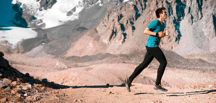 A young man runs along a trail in the snow-capped mountains, doing outdoor workout. The runner warms up, prepares for the race, leads a healthy active lifestyle.