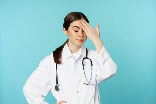 Portrait of annoyed, tired woman doctor, facepalm, roll eyes frustrated, bothered by smth stupid, standing in white coat over torquoise background.