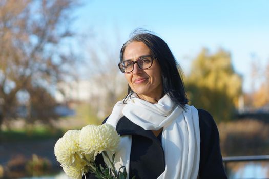 A beautiful middle-aged woman getting grey-haired in a dark coat in a spring town with a bouquet of flowers