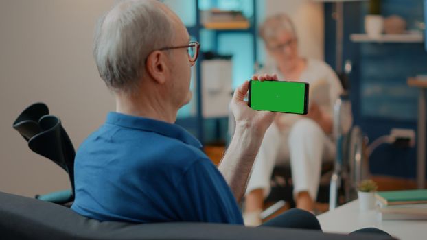 Elder man using horizontal green screen on mobile phone, having crutches on sofa. Retired person holding chroma key template with isolated copy space and blank mock up background on device.