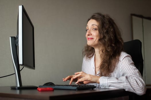 Young female office manager in white shirt and curly hair is sitting at the table and typing using keyboard, routine work, freelance.