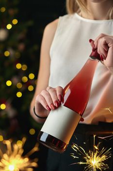 Bottle of rose wine in hands of smartly dressed young woman against blurred background of Christmas decorations with bright sparklers. New Years celebration concept