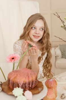 A girl with long hair in a light dress is sitting at the Easter table with cakes, spring flowers and quail eggs. Happy Easter celebration