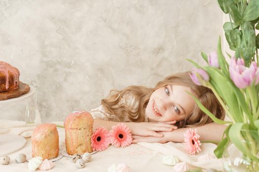 A girl with long hair in a light dress is sitting at the Easter table with cakes, spring flowers and quail eggs. Happy Easter celebration