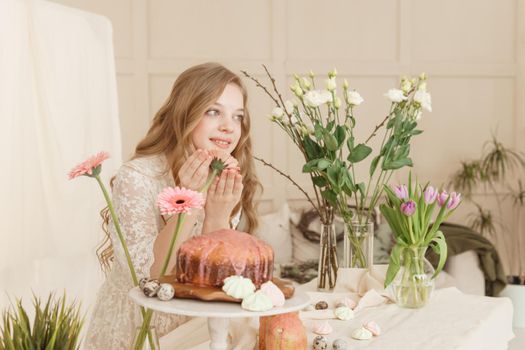 A girl with long hair in a light dress is sitting at the Easter table with cakes, spring flowers and quail eggs. Happy Easter celebration
