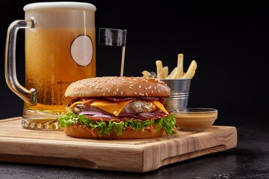 Appetizing cheeseburger with pork and veal patty, fried bacon, fresh red onion, tomatoes and lettuce in sesame bun served with glass of foamy amber beer, crispy fries and aioli sauce on wooden board