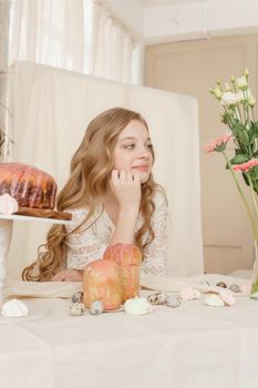 A girl with long hair in a light dress is sitting at the Easter table with cakes, spring flowers and quail eggs. Happy Easter celebration